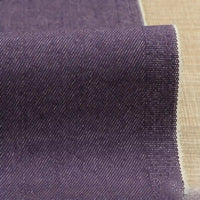 A close-up of Longan Craft’s 9.5oz Stretch Denim Fabric by the yard in dark purple, featuring a subtle diagonal weave—an ideal solid cotton denim textile for jeans—shown on a light beige textured surface.