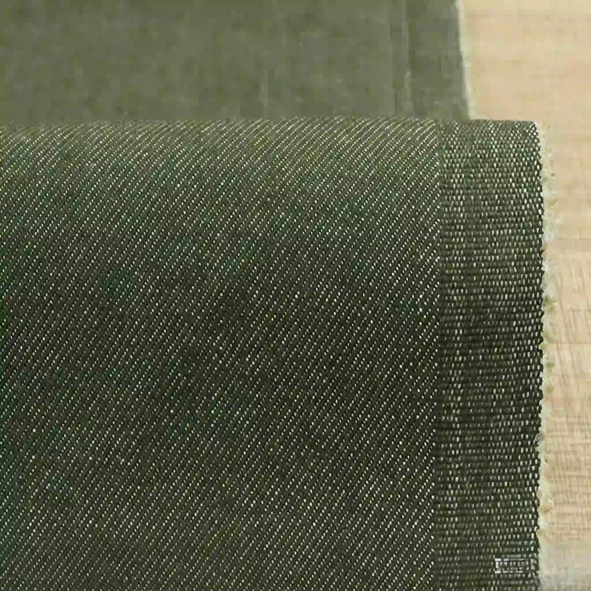 A close-up of Longan Craft’s 9.5oz Stretch Denim Fabric By The Yard shows its dark green, medium-weight cotton material with a subtle diagonal weave and neatly finished selvage on a light wood surface—perfect for jeans and denim projects.