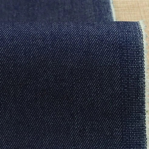 A close-up of Longan Craft's 9.5oz Stretch Denim Fabric By The Yard in solid dark blue cotton, showing diagonal weaving patterns and a slightly frayed edge, displayed on a light beige surface—ideal material for jeans or crafts.