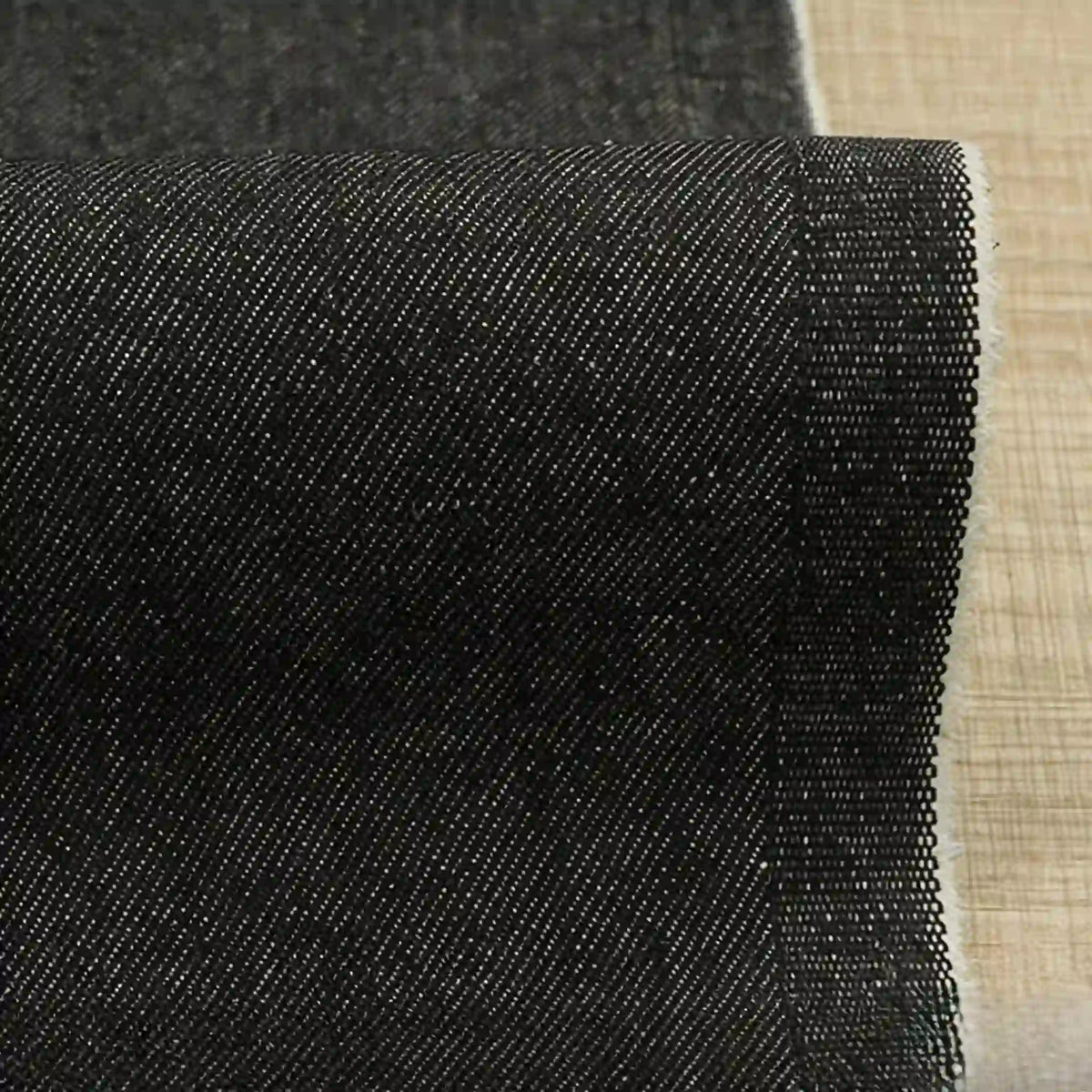 A close-up of Longan Craft’s 9.5oz Stretch Denim Fabric By The Yard in solid black shows its diagonal twill weave and raw edge, ideal cotton denim textile for jeans, displayed on a beige surface.