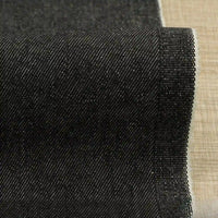 A close-up of Longan Craft’s 9.5oz Stretch Denim Fabric By The Yard in solid black shows its diagonal twill weave and raw edge, ideal cotton denim textile for jeans, displayed on a beige surface.