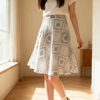 A woman stands indoors in sunlight beside a window, wearing a white top and a knee-length skirt made from the Longan Craft Women A-line Midi Skirt Sewing Pattern—available as a free PDF skirt pattern with gray geometric and floral designs.