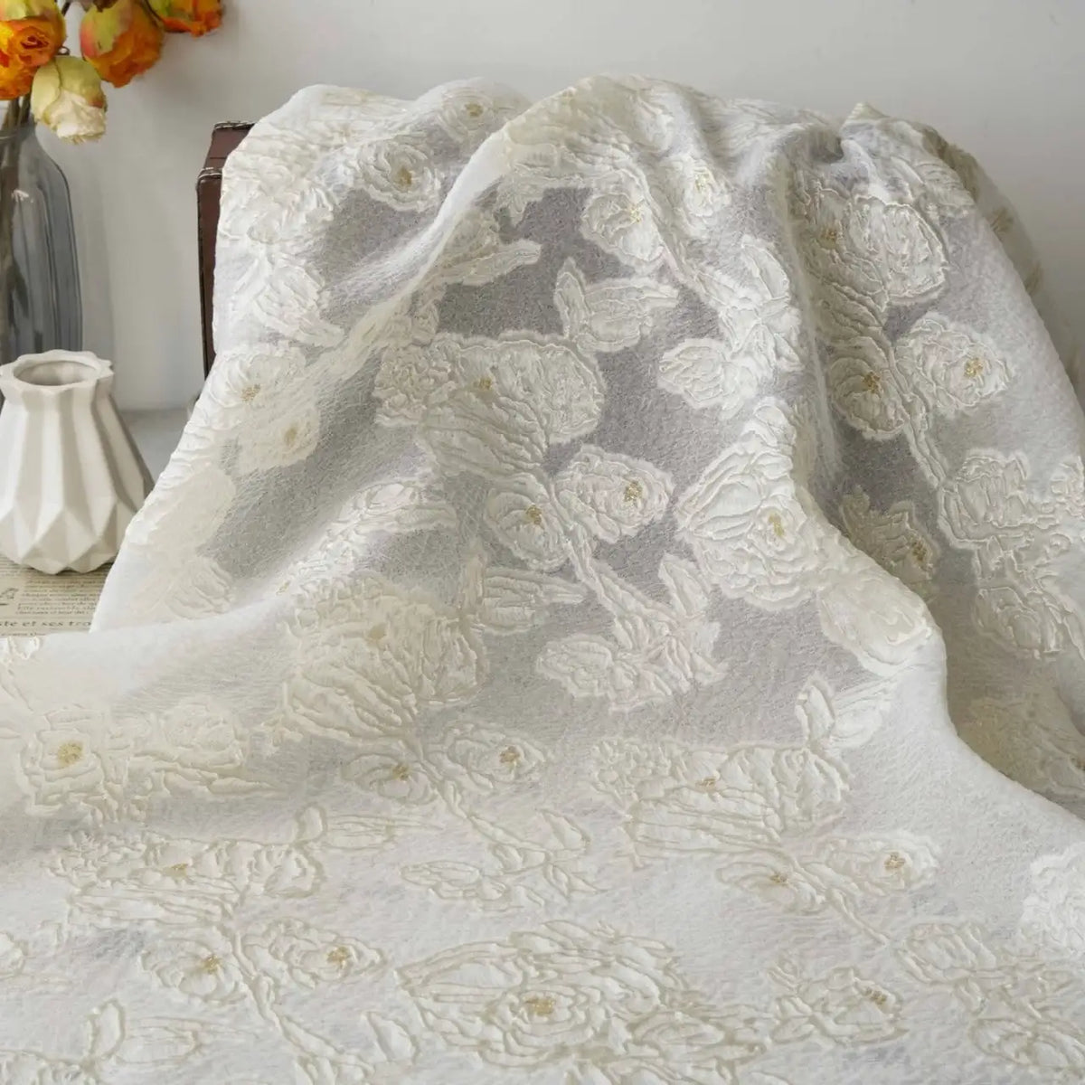 Longan Craft's Apricot Rose Embossed Organza Fabric By The Yard, featuring 3D white floral jacquard embroidery, is elegantly draped with a white vase and a glass vase of orange flowers providing delicate contrast in the background.