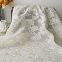 Longan Craft's Apricot Rose Embossed Organza Fabric By The Yard, featuring 3D white floral jacquard embroidery, is elegantly draped with a white vase and a glass vase of orange flowers providing delicate contrast in the background.