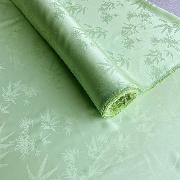 A roll of Longan Craft’s Bamboo Jacquard Mulberry Silk Fabric By The Yard, featuring a subtle leafy pattern and smooth, slightly shiny texture in light green, is partially unrolled on a flat surface.