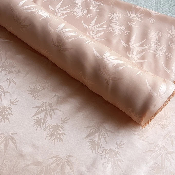 A roll of Longan Craft Bamboo Jacquard Mulberry Silk Fabric by the yard, with a smooth, silky surface and subtle leaf pattern in light peach, is spread flat to showcase its elegant, cozy design—ideal for lining or colorful rayon silk projects.