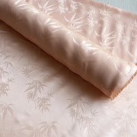 A roll of Longan Craft Bamboo Jacquard Mulberry Silk Fabric by the yard, with a smooth, silky surface and subtle leaf pattern in light peach, is spread flat to showcase its elegant, cozy design—ideal for lining or colorful rayon silk projects.