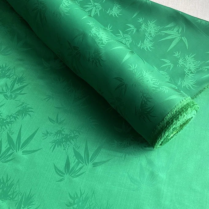 A roll of Longan Craft Bamboo Jacquard Mulberry Silk Fabric, featuring a subtle, glossy botanical leaf pattern in green, is spread out flat—ideal as colorful rayon silk or cozy silk lining fabric by the yard.