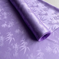 A partially unrolled roll of Longan Craft Bamboo Jacquard Mulberry Silk Fabric By The Yard, featuring a light purple color and glossy bamboo leaf pattern, perfect as a colorful rayon silk lining fabric.