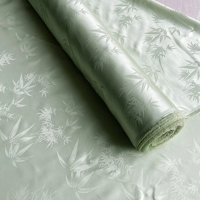 A roll of Bamboo Jacquard Mulberry Silk Fabric by Longan Craft, featuring a subtle woven pattern in pale green, is partially unrolled on a flat surface.