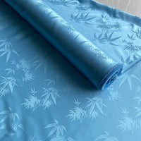 A roll of Bamboo Jacquard Mulberry Silk Fabric by Longan Craft in light blue is partially unrolled, revealing its subtle, glossy leaf pattern—perfect as a colorful silk lining fabric or for cozy sewing projects.