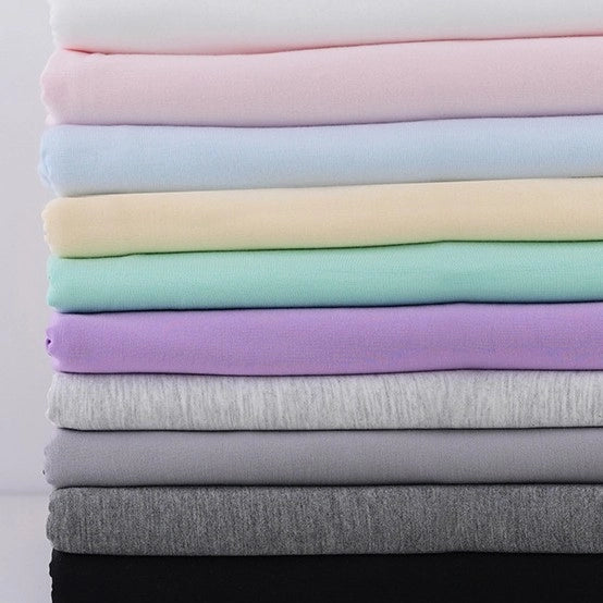 A stack of Longan Craft's Bamboo Jersey Fabric By The Yard in white, pink, yellow, mint green, lavender, light gray, dark gray, and black—soft plain color material perfect for loungewear and eco-friendly fabric projects.