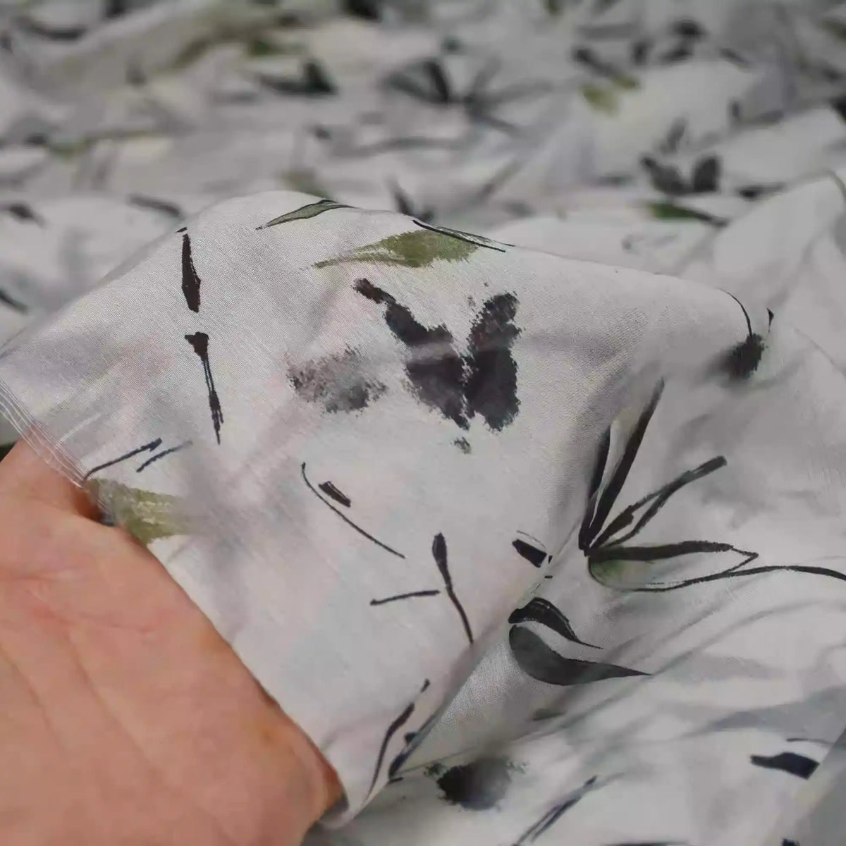 A hand holds Longan Craft's Bamboo Printed Nylon Viscose Fabric by the yard, a lightweight, eco-friendly Chinoiserie viscose with a sheer white base and delicate black and green floral-leaf print, perfect for premium dressmaking.