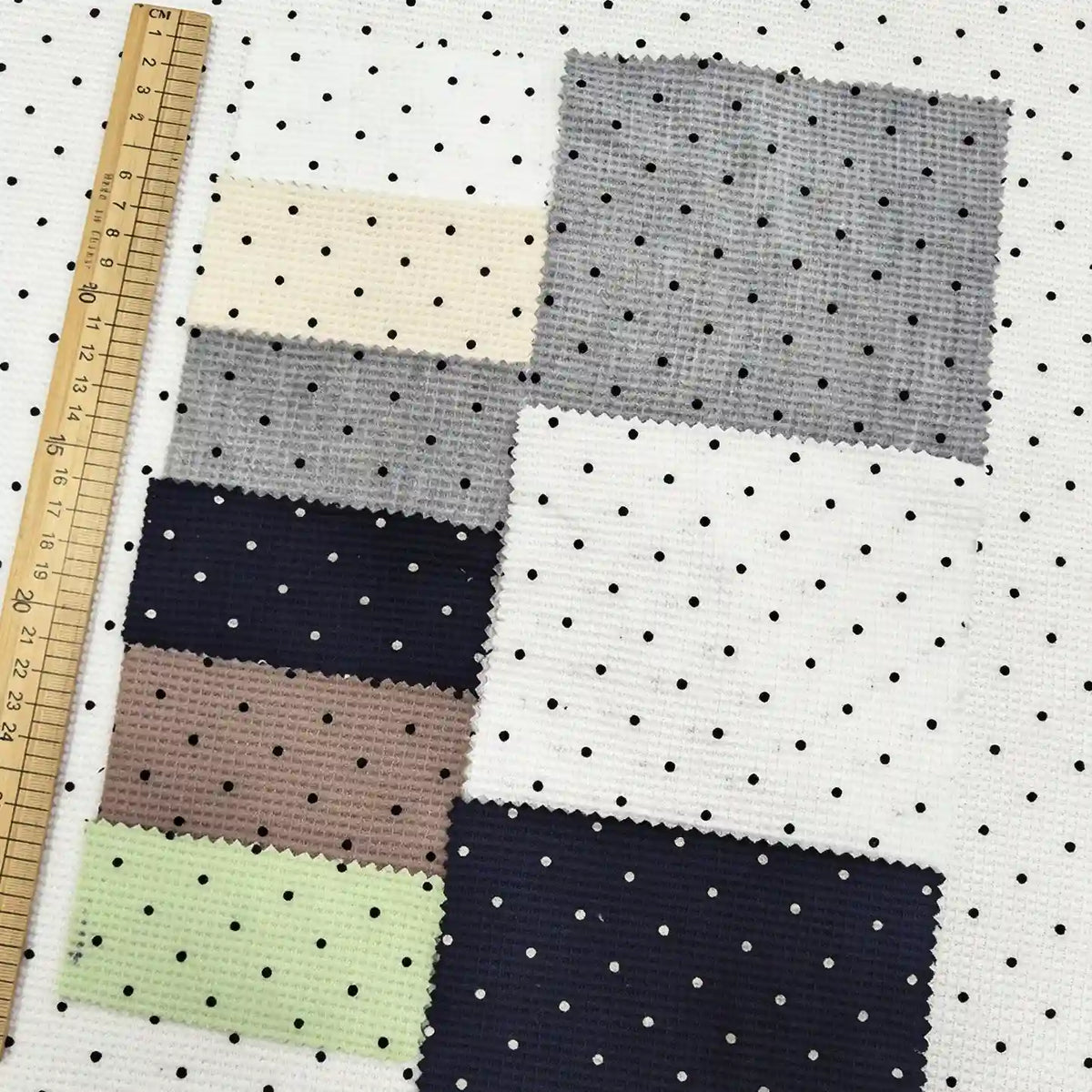 A collection of square fabric swatches, including Black Mini Polka Dot Print Waffle Fabric By The Yard, laid over a white polka dot T-shirt fabric next to a wooden ruler.