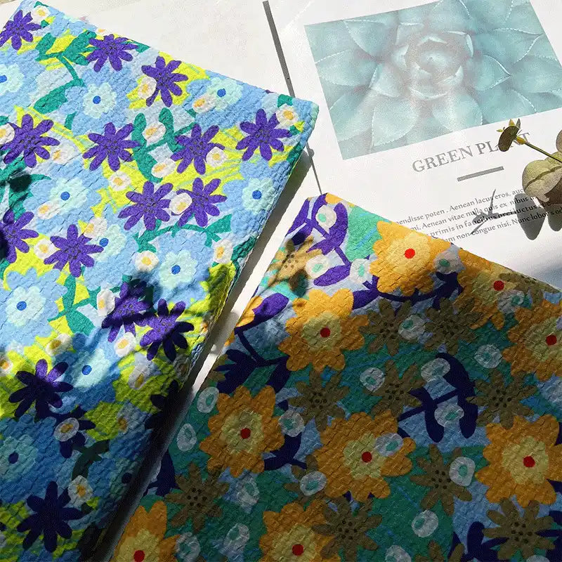 Two botanical printed seersucker cloths from Longan Craft, made of breathable puckered cotton fabric and ideal for spring and summer skirts, are displayed on a sunlit surface beside a paper featuring a photo of succulents and the words GREEN PLANTS. Leaf shadows gently fall across the scene.