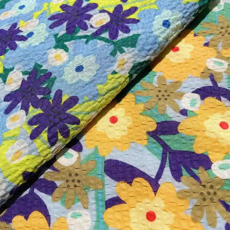 Crafted by Longan Craft, the Botanical Printed Seersucker Fabric By The Yard features two pieces of puckered cotton seersucker cloth adorned with floral patterns. One side displays blue and purple flowers, while the other showcases yellow, brown, and purple blooms set against a blue background. This breathable seersucker material offers comfort and charm—perfect for your spring and summer creative projects such as skirts.
