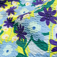 Close-up of Longan Craft’s Botanical Printed Seersucker Fabric By The Yard, featuring a floral puckered cotton surface adorned with blue, purple, and yellow flowers accented by green leaves on a light background. The fabric showcases visible raised dots reminiscent of classic seersucker texture, making it an ideal material for spring and summer skirts.