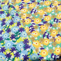 Two pieces of Longan Craft’s Botanical Printed Seersucker Fabric: one side displays a delightful blue and purple floral pattern on a light, breathable seersucker cotton background, while the other features charming yellow and white flowers with red centers set against a greenish backdrop—perfect for spring and summer projects with this textured puckered fabric.