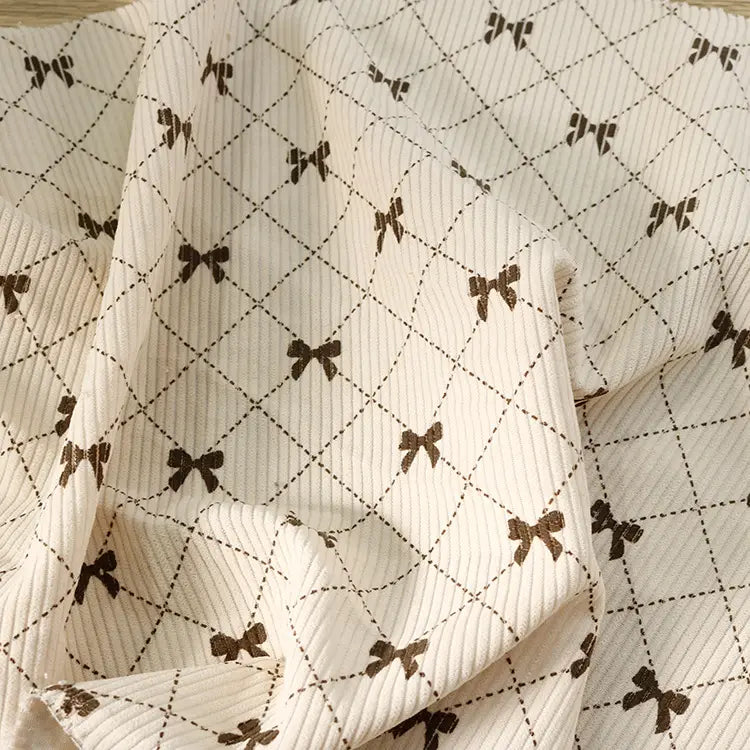 Introducing the Bow Plaid Print Corduroy Fabric By The Yard from Longan craft—a heavyweight, soft, and durable corduroy clothing fabric. This cream-colored material features a ribbed texture with a diamond grid pattern adorned with delicate brown bow designs at each intersection. The slightly rumpled finish creates gentle folds and subtle shadows, adding depth to its classic checked print.