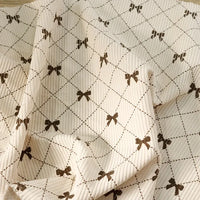 Introducing the Bow Plaid Print Corduroy Fabric By The Yard from Longan craft—a heavyweight, soft, and durable corduroy clothing fabric. This cream-colored material features a ribbed texture with a diamond grid pattern adorned with delicate brown bow designs at each intersection. The slightly rumpled finish creates gentle folds and subtle shadows, adding depth to its classic checked print.