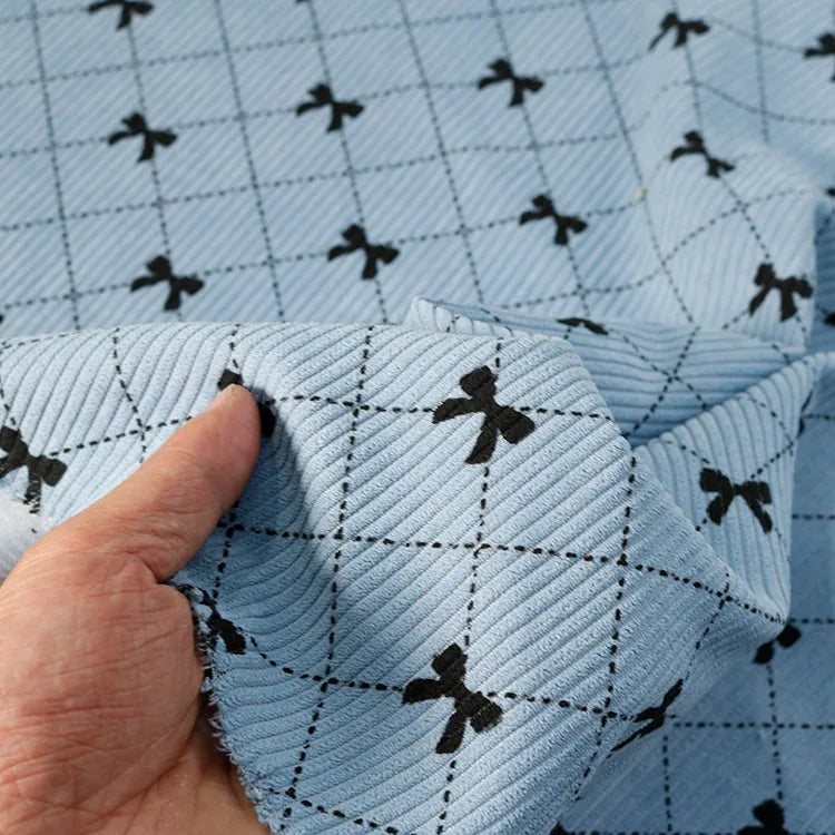 A hand displays the Bow Plaid Print Corduroy Fabric by Longan craft, featuring light blue heavyweight corduroy adorned with a black grid and small black bows at each intersection—an eye-catching, soft and durable checked fabric perfect for clothing projects.