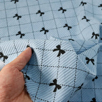 A hand displays the Bow Plaid Print Corduroy Fabric by Longan craft, featuring light blue heavyweight corduroy adorned with a black grid and small black bows at each intersection—an eye-catching, soft and durable checked fabric perfect for clothing projects.