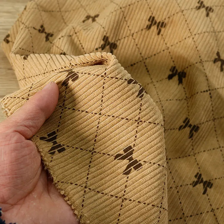 A hand holds a piece of Bow Plaid Print Corduroy Fabric By The Yard from Longan craft, featuring a light brown, heavyweight checked print with diamond grid patterns and small dark motifs at the intersections, reminiscent of printed corduroy material.
