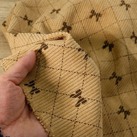 A hand holds a piece of Bow Plaid Print Corduroy Fabric By The Yard from Longan craft, featuring a light brown, heavyweight checked print with diamond grid patterns and small dark motifs at the intersections, reminiscent of printed corduroy material.