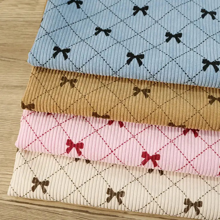 Four folded pieces of Longan craft Bow Plaid Print Corduroy Fabric By The Yard in blue, tan, pink, and cream—each showcasing a diamond grid plaid pattern with small bow designs—are stacked neatly on a wooden surface. This heavyweight checked printed corduroy material is both soft and durable, ideal for clothing projects.