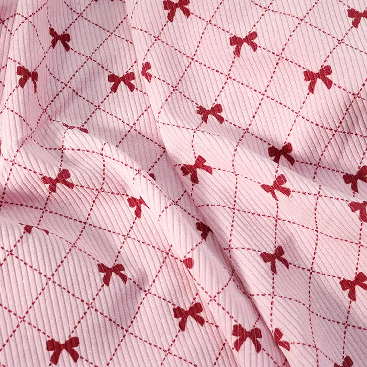 Bow Plaid Print Corduroy Fabric By The Yard from Longan craft is a heavyweight, soft, and durable corduroy material featuring a textured pink surface adorned with a repeating pattern of red bows and intersecting dashed red lines that form a charming diamond grid. The checked printed corduroy is gently folded, highlighting subtle shadows and its unique bow plaid design—perfect for clothing and crafts.