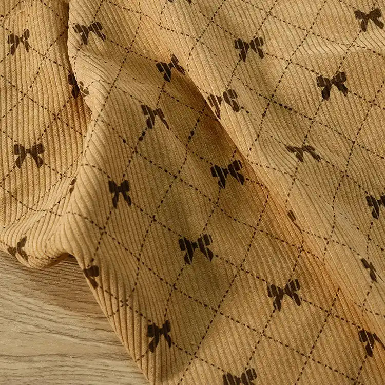 Longan craft’s Bow Plaid Print Corduroy Fabric By The Yard—a heavyweight, soft, and durable checked printed corduroy material—features a beige background with a brown diamond grid pattern and small bow designs at each intersection, elegantly draped on a light wood surface.