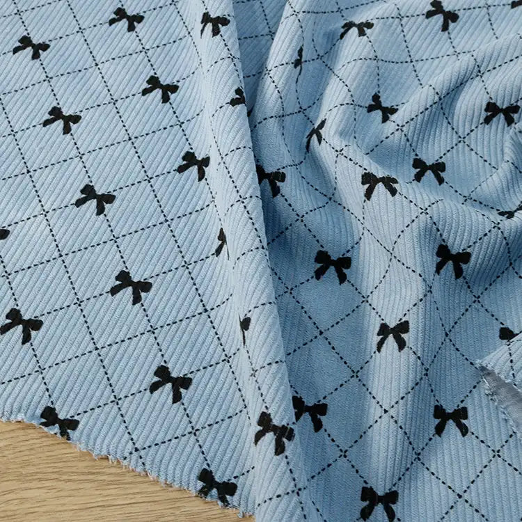 Longan craft Bow Plaid Print Corduroy Fabric By The Yard, a heavyweight checked printed corduroy material, features a soft and durable ribbed texture in light blue with a grid of thin dark lines and small black bows at each intersection, displayed on a wooden surface.
