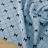 Longan craft Bow Plaid Print Corduroy Fabric By The Yard, a heavyweight checked printed corduroy material, features a soft and durable ribbed texture in light blue with a grid of thin dark lines and small black bows at each intersection, displayed on a wooden surface.