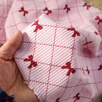 A hand holds a piece of Longan craft Bow Plaid Print Corduroy Fabric By The Yard, a soft and durable heavyweight checked corduroy featuring a textured pink surface adorned with a red grid pattern and small red bows at each intersection.