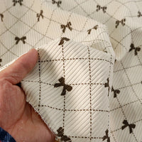 A hand holds a piece of cream-colored, ribbed fabric featuring a brown grid pattern with small bows at the intersections, highlighting Longan craft’s Bow Plaid Print Corduroy Fabric By The Yard—a soft and durable heavyweight checked printed corduroy material perfect for clothing projects.