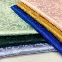 Stacks of Longan Craft's Brocade Damask Fabric By The Yard in mint green, pink, gold, royal blue, dark green, and lavender—each with a glossy floral vintage jacquard pattern—perfect for evening gowns or damask curtain material.