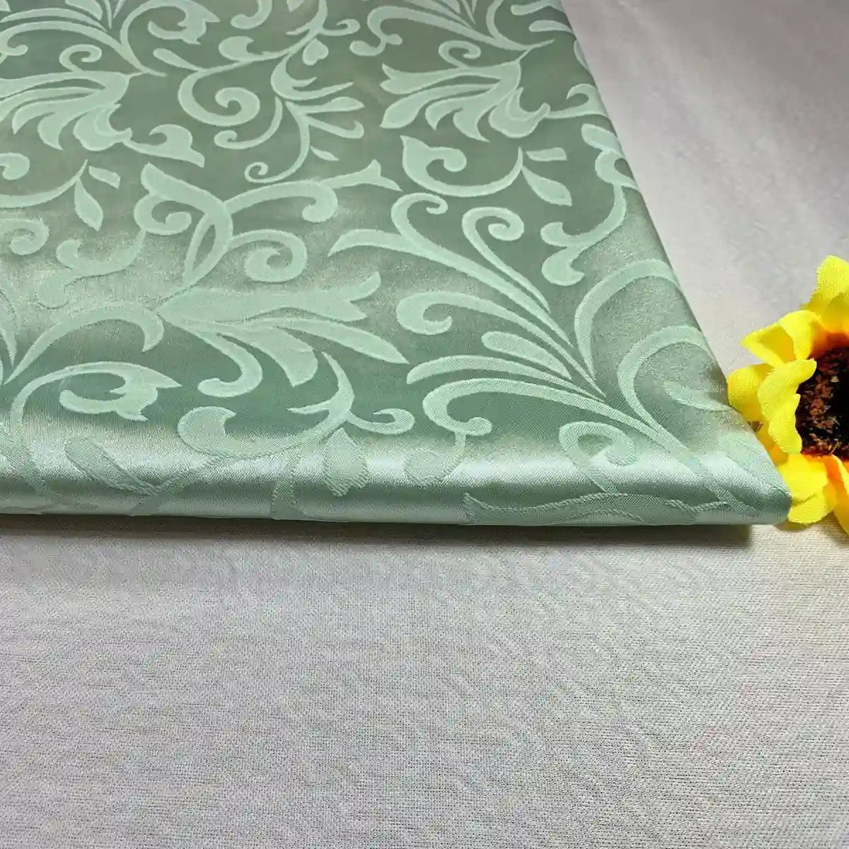 A folded piece of light green Brocade Damask Fabric By The Yard from Longan Craft, featuring a raised floral pattern, lies on a plain surface next to a yellow artificial sunflower.