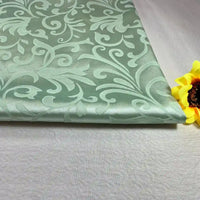 A folded piece of light green Brocade Damask Fabric By The Yard from Longan Craft, featuring a raised floral pattern, lies on a plain surface next to a yellow artificial sunflower.
