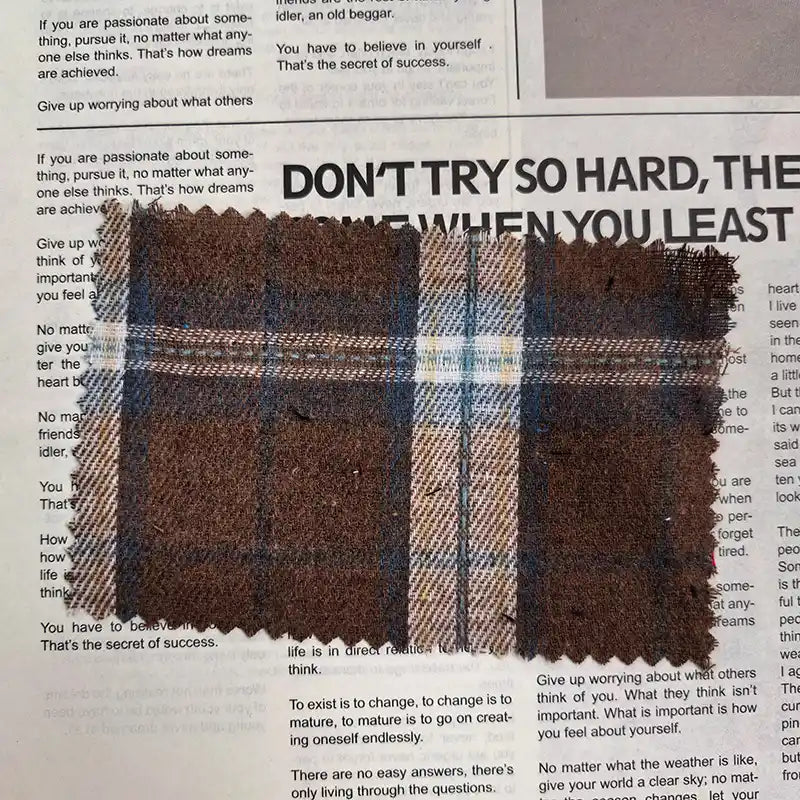 A swatch of Longan Craft’s Brown Plaid Cotton Flannel Fabric By The Yard—featuring brown, blue, and white plaid on lightweight polyester brushed flannel—is displayed atop a newspaper with visible black text and headlines.