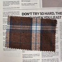 A swatch of Longan Craft’s Brown Plaid Cotton Flannel Fabric By The Yard—featuring brown, blue, and white plaid on lightweight polyester brushed flannel—is displayed atop a newspaper with visible black text and headlines.