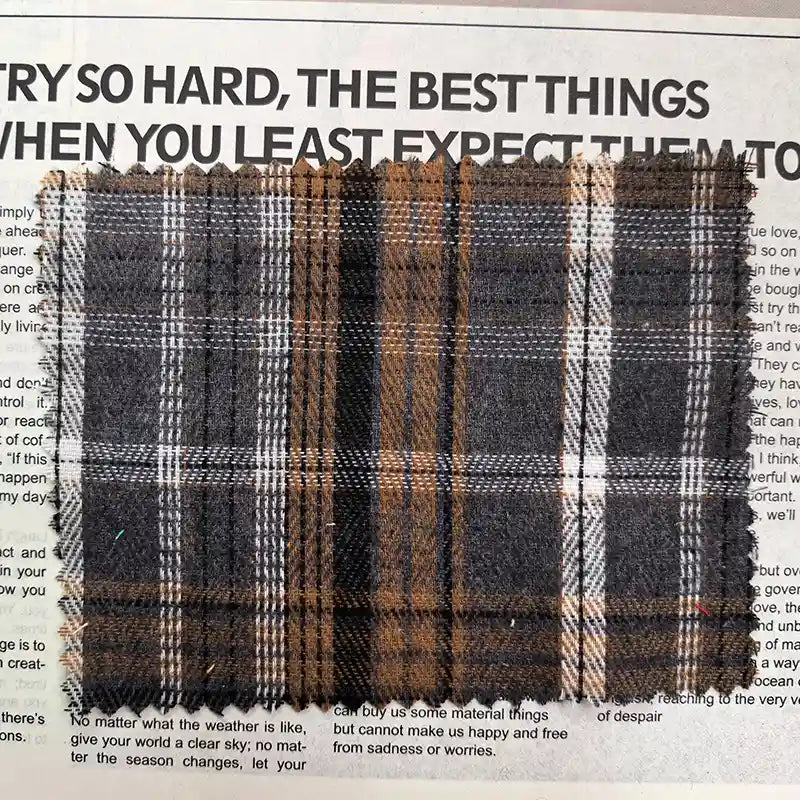 A piece of Longan Craft’s Brown Plaid Cotton Flannel Fabric By The Yard—featuring a retro plaid pattern with gray, black, and white stripes on brown lightweight polyester brushed flannel—is displayed atop newspaper text.