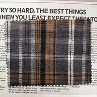A piece of Longan Craft’s Brown Plaid Cotton Flannel Fabric By The Yard—featuring a retro plaid pattern with gray, black, and white stripes on brown lightweight polyester brushed flannel—is displayed atop newspaper text.