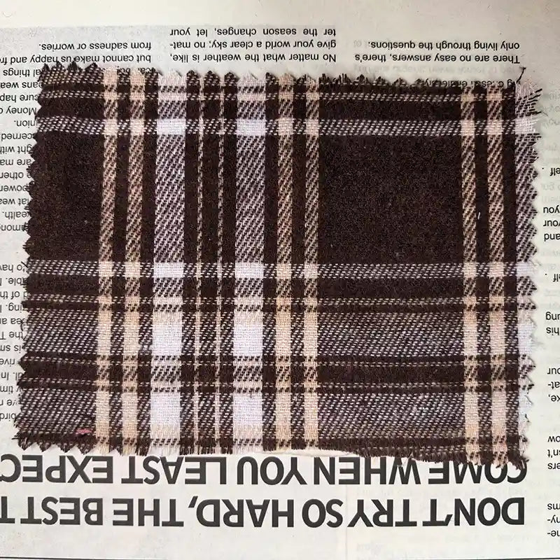 A swatch of Longan Craft’s Brown Plaid Cotton Flannel Fabric By The Yard— a lightweight polyester brushed flannel material ideal for retro casual pants— is placed on a newspaper, with bold, upside-down black text partially visible along the bottom edge.