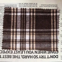 A swatch of Longan Craft’s Brown Plaid Cotton Flannel Fabric By The Yard— a lightweight polyester brushed flannel material ideal for retro casual pants— is placed on a newspaper, with bold, upside-down black text partially visible along the bottom edge.