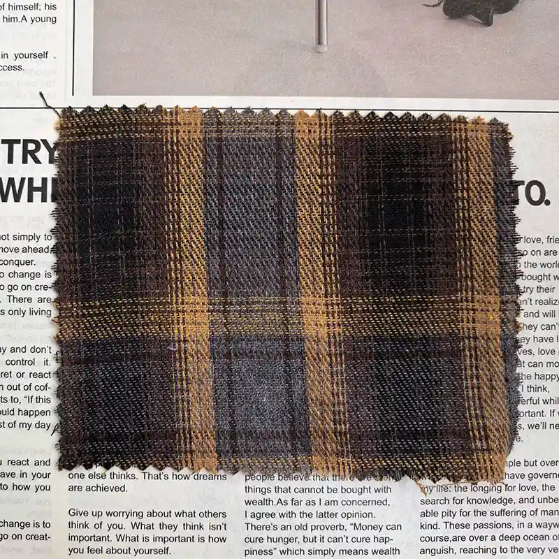 A rectangular swatch of Longan Craft’s Brown Plaid Cotton Flannel Fabric By The Yard—a lightweight, polyester brushed flannel material ideal for casual pants—is displayed on top of a newspaper page with visible black text.
