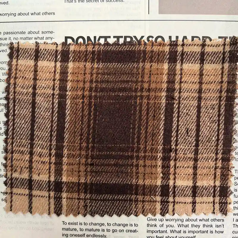 A square piece of Brown Plaid Cotton Flannel Fabric By The Yard from Longan Craft is placed atop a magazine or newspaper page. This lightweight polyester brushed flannel material showcases a retro brown and beige plaid design, bringing warmth and texture to the scene.