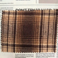 A square piece of Brown Plaid Cotton Flannel Fabric By The Yard from Longan Craft is placed atop a magazine or newspaper page. This lightweight polyester brushed flannel material showcases a retro brown and beige plaid design, bringing warmth and texture to the scene.
