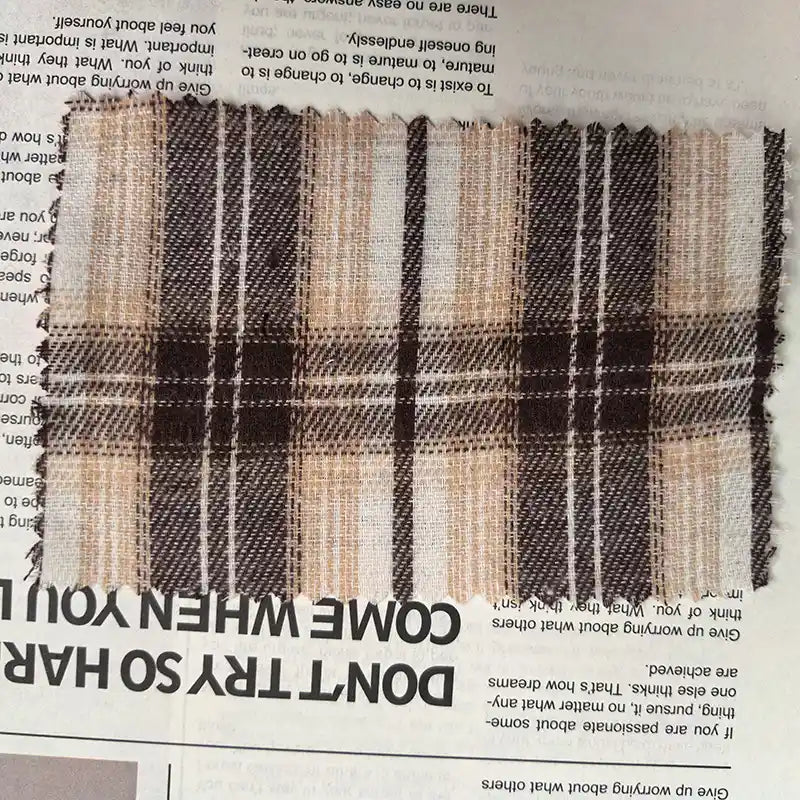 A rectangular swatch of Longan Craft’s Brown Plaid Cotton Flannel Fabric By The Yard, a lightweight polyester brushed flannel featuring a brown, beige, and white plaid pattern and zigzag-cut frayed edges, lies on a newspaper.