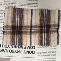 A rectangular swatch of Longan Craft’s Brown Plaid Cotton Flannel Fabric By The Yard, a lightweight polyester brushed flannel featuring a brown, beige, and white plaid pattern and zigzag-cut frayed edges, lies on a newspaper.