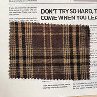 A rectangular swatch of Longan Craft’s Brown Plaid Cotton Flannel Fabric By The Yard, featuring a lightweight polyester brushed flannel material with beige and dark lines, rests atop a printed page with black text.